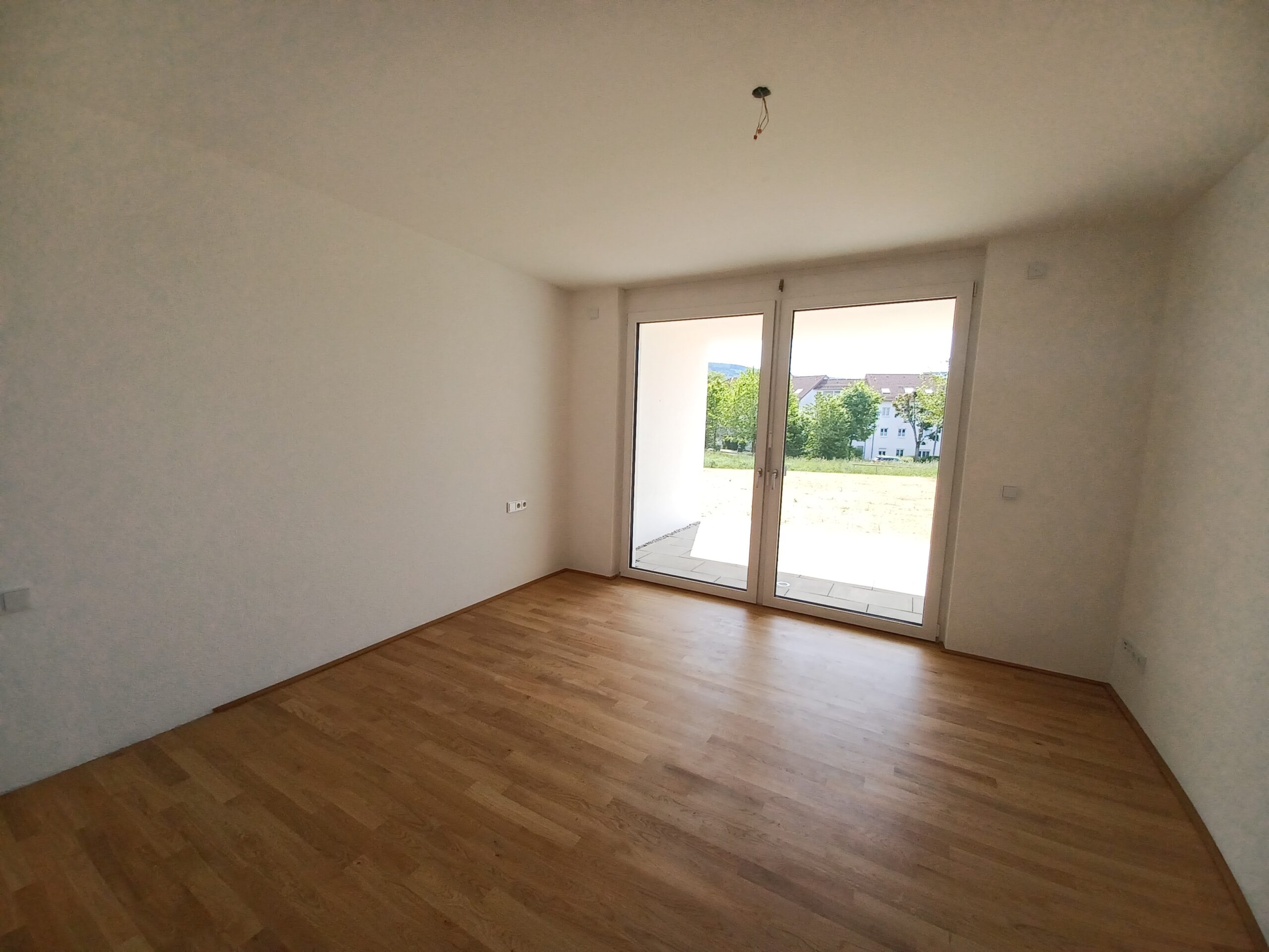 Innenansicht eines hellen Zimmers mit Echtholzboden, weiß gestrichenen Wänden und großen bodentiefen Fenstertüren, die auf eine gepflasterte Terrasse und eine begrünte Außenfläche führen.