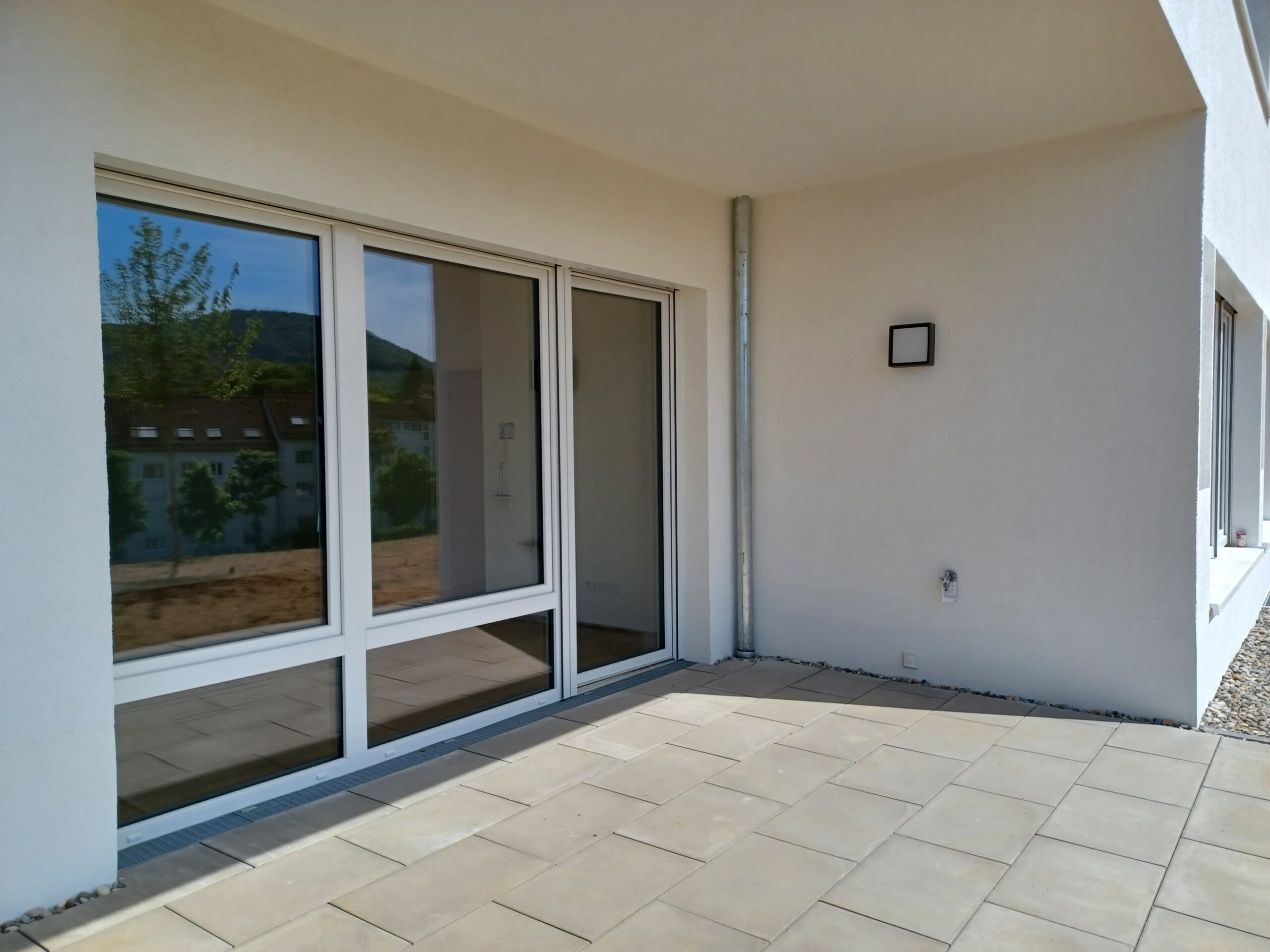 Foto einer überdachten Terrasse mit hellen Steinplatten, angrenzend an eine weiß verputzte Hausfassade. Eine große Fensterfront mit Terrassentür bildet den Zugang zur Wohnung. Rechts an der Wand befindet sich eine Außenleuchte und ein Wasseranschluss. In der Spiegelung der Fenster sind Bäume und ein gegenüberliegendes Wohnhaus sichtbar.