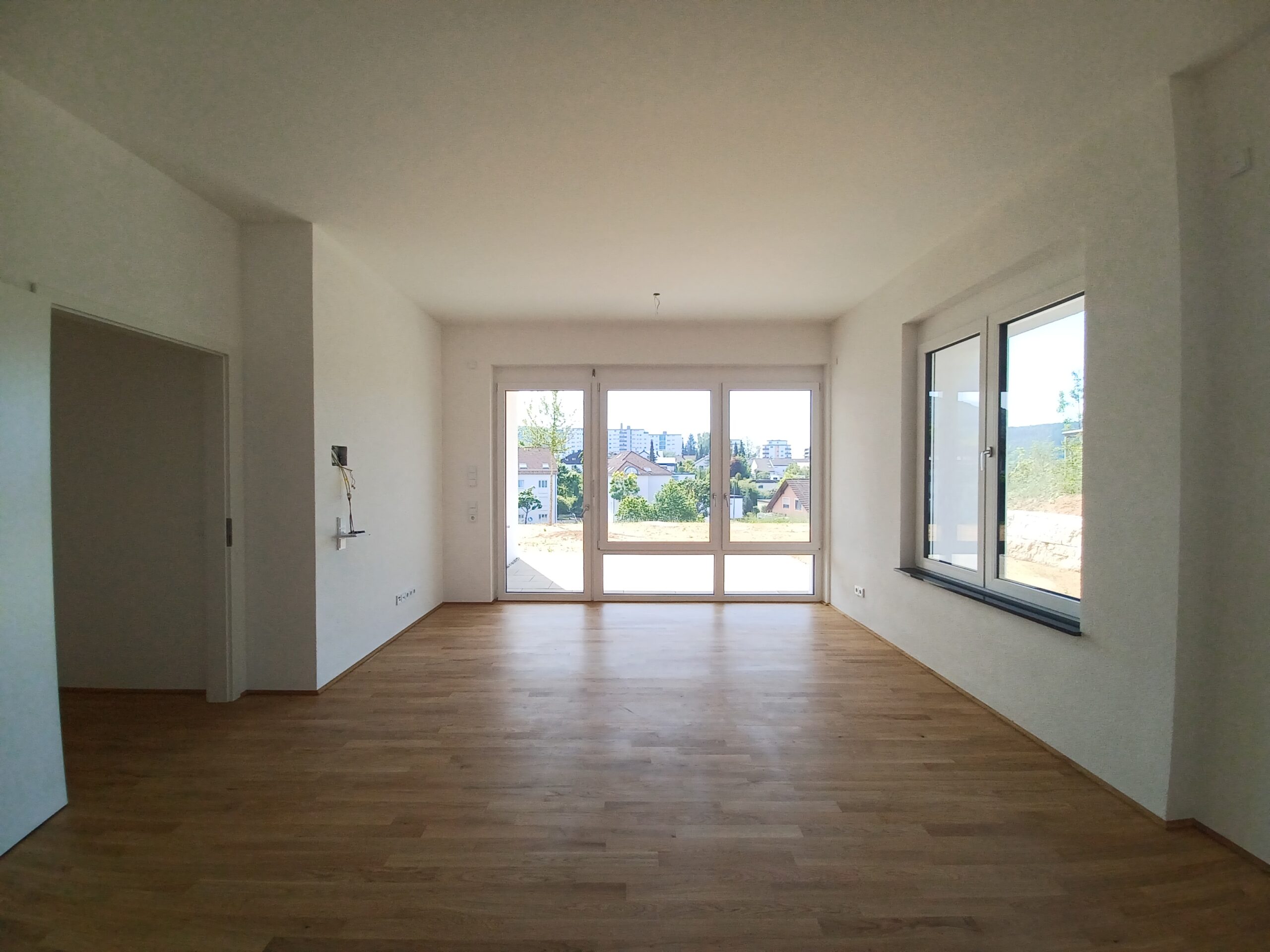 Foto eines leeren Wohnzimmers mit Holzfußboden, weißen Wänden und großen bodentiefen Fenstern auf der Stirnseite. Die Fenster und Terrassentür bieten Ausblick auf eine grüne Umgebung mit Wohnhäusern und Stadt im Hintergrund. Rechts befindet sich ein weiteres großes Fenster, links ein offener Türrahmen zu einem angrenzenden Raum.