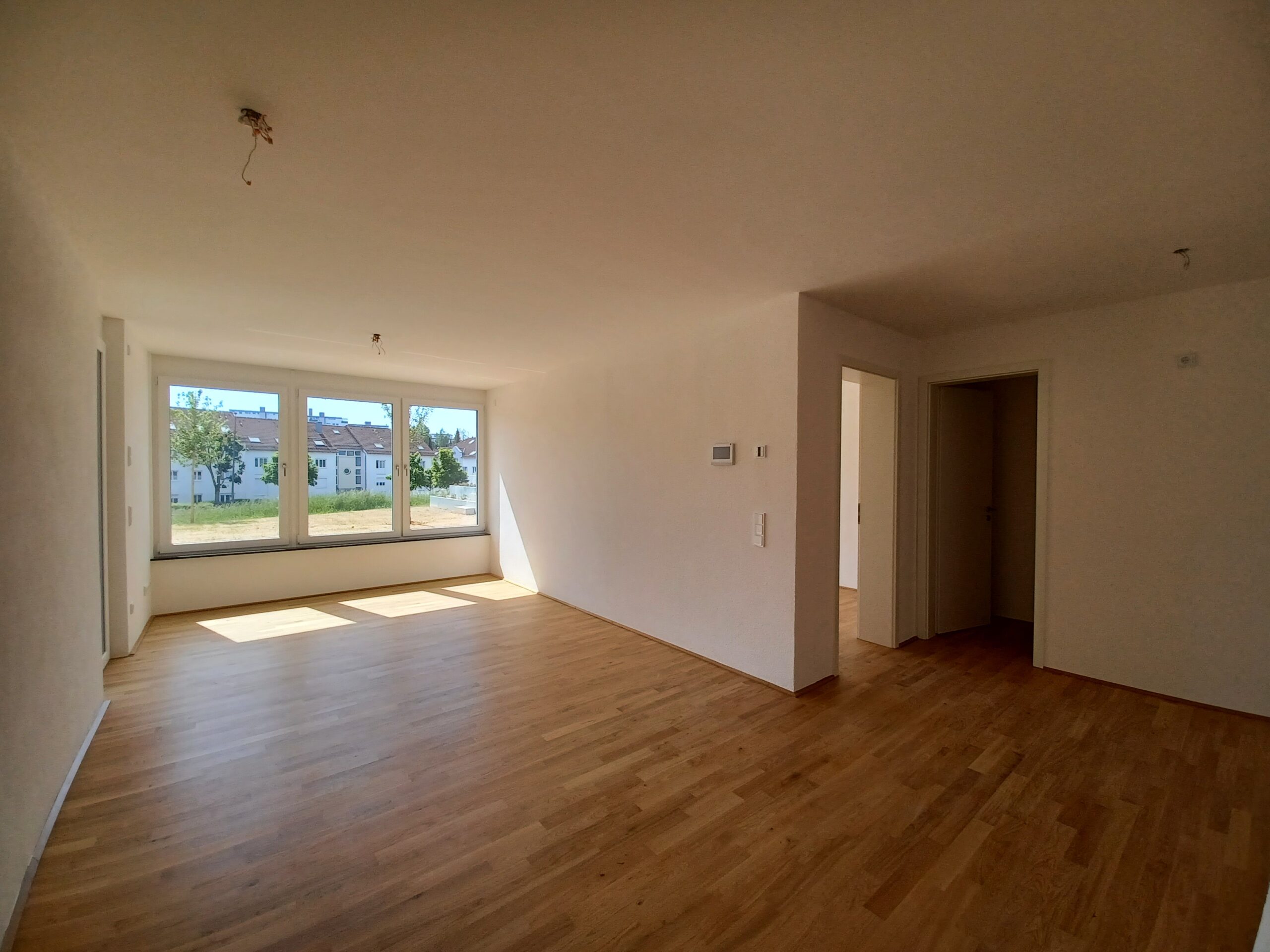 Innenaufnahme eines hellen Wohnraums mit bodentiefem Fenster mit Blick auf eine Grünfläche und angrenzendem Zugang zu zwei weiteren Zimmern über einen Flur. Der Raum verfügt über einen Echtholzboden und weiße Wände.