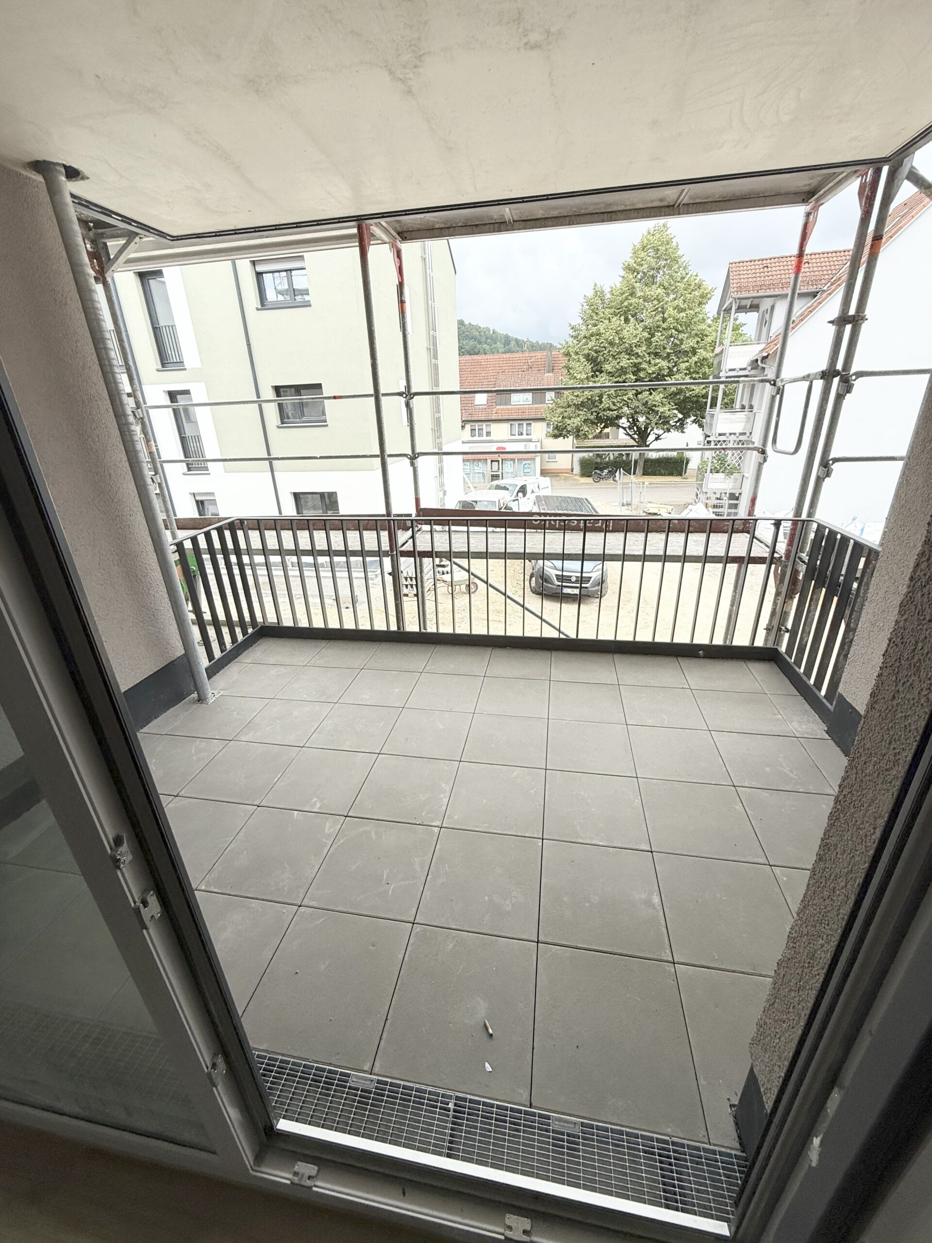 Innenaufnahme eines hellen Wohnraums mit bodentiefem Fenster mit Blick auf eine Grünfläche und angrenzendem Zugang zu zwei weiteren Zimmern über einen Flur. Der Raum verfügt über einen Echtholzboden und weiße Wände.