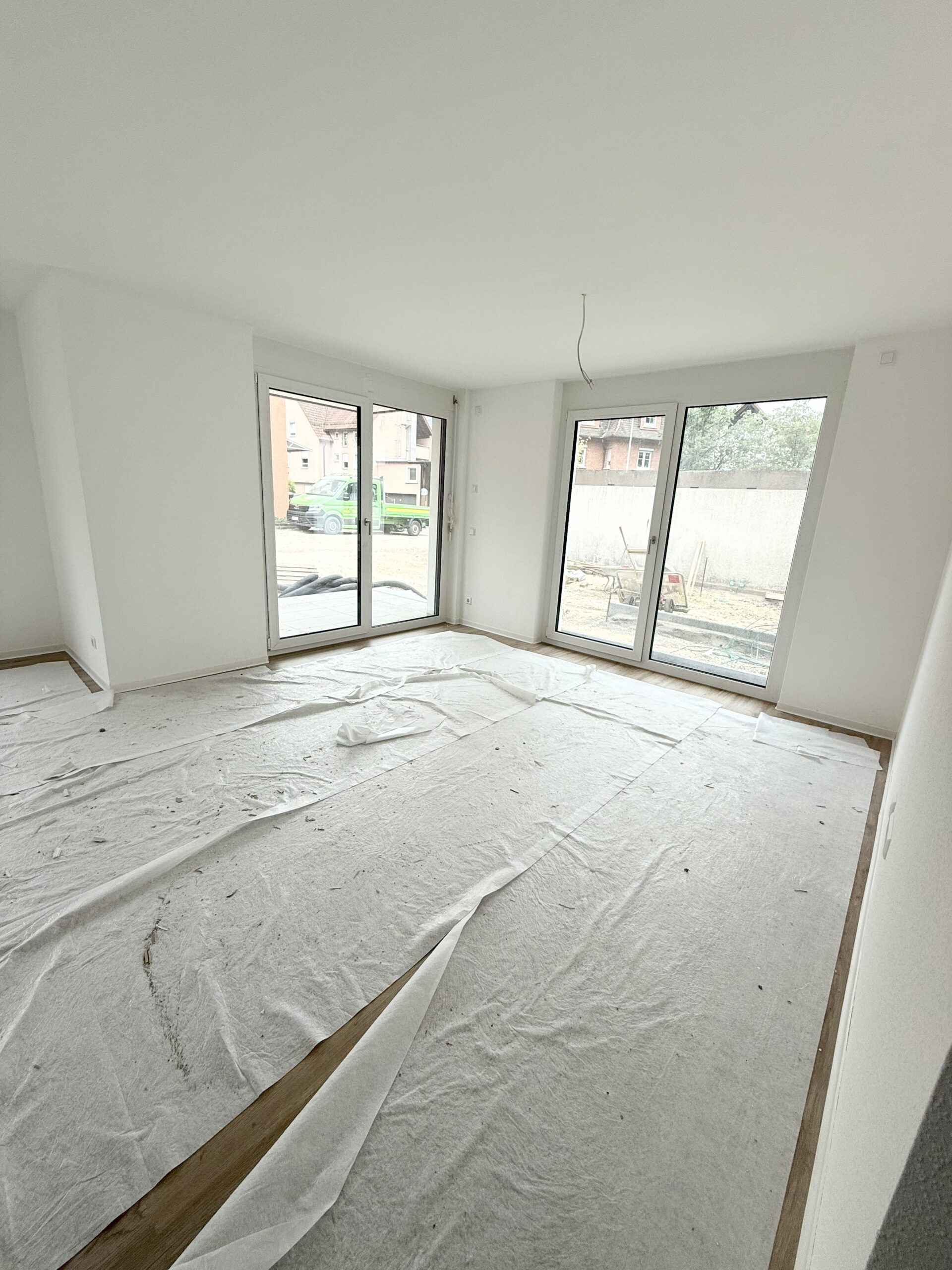 Innenaufnahme eines hellen Wohnraums mit bodentiefem Fenster mit Blick auf eine Grünfläche und angrenzendem Zugang zu zwei weiteren Zimmern über einen Flur. Der Raum verfügt über einen Echtholzboden und weiße Wände.